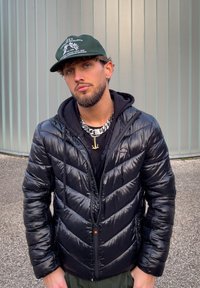 Jeune homme portant une casquette noire, une doudoune noire, un sweat à capuche, une chaîne en argent et un pendentif en forme d'ancre, debout à l'extérieur près d'un mur à panneaux métalliques.