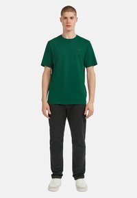 Zaļa īsām piedurknēm T-krekls, izgatavots no kokvilnas, ar apaļu kakla izgriezumu un mazu logotipa detaļu kreisajā krūtī. Valkāts ar melniem biksēm un baltām kurpēm.