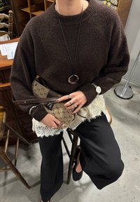 Bruine gebreide trui met verlaagde schouders, gecombineerd met gelaagde kanten en op maat gemaakte zwarte broek. Accessoires omvatten een handtas met patroon en ringen.