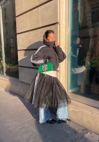 Femme en doudoune noire avec rayures blanches, jupe en tulle noire portée sur un jean bleu, chaussures noires, tenant un sac à main vert, debout près d'une fenêtre de bâtiment.