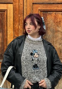 Grigio maglione lavorato a maglia con un design ricamato di Minnie Mouse, abbinato a una giacca di pelle nera e a un dolcevita bianco sotto.