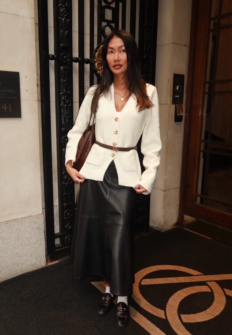 Femme portant un cardigan blanc boutonné avec ceinture marron, jupe midi en cuir noir, mocassins noirs avec chaussettes blanches, et sac à bandoulière marron, debout à l'intérieur.