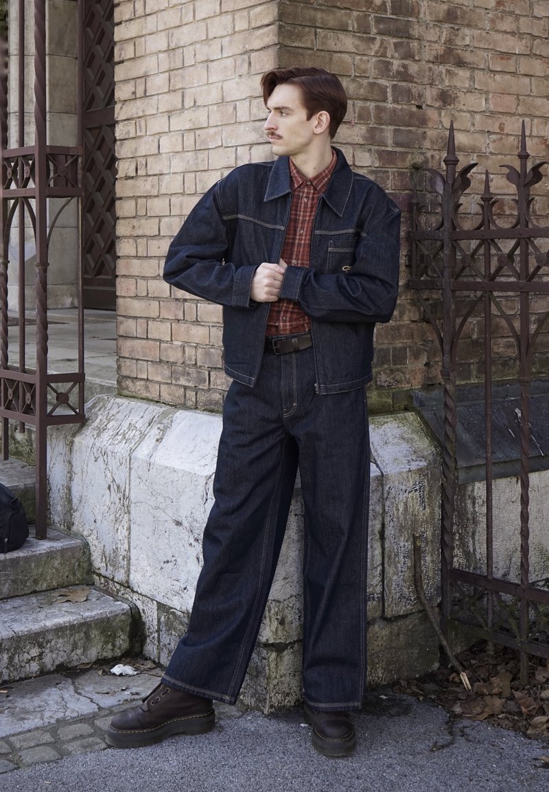Hombre con chaqueta y pantalones de mezclilla oscura y camisa de cuadros rojos, de pie contra una pared de ladrillos cerca de una verja metálica, mirando hacia la izquierda.