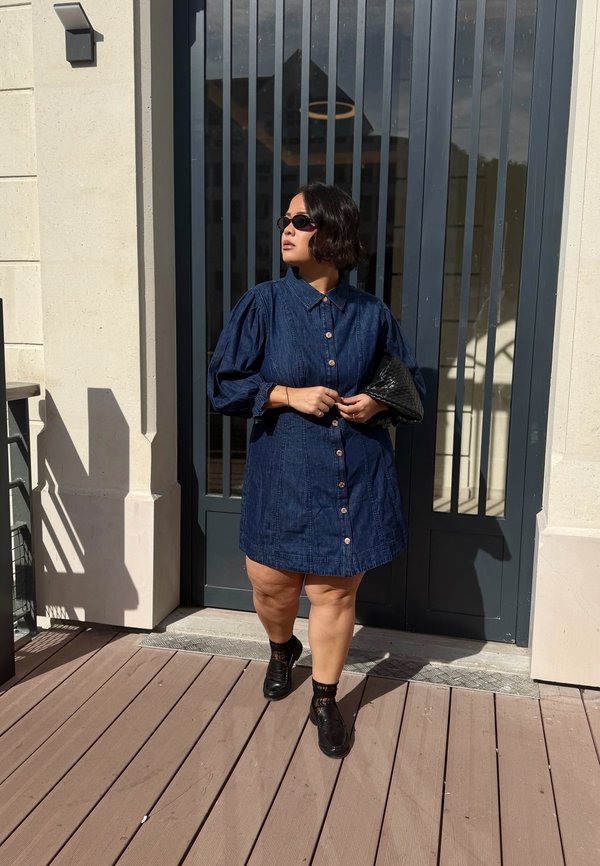 Femme portant une robe en denim bleue avec des manches bouffantes et des lunettes de soleil se tient devant une porte en verre sombre en tenant une pochette noire.
