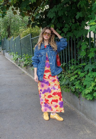 Chaqueta de denim sobre un maxi vestido de tie-dye vibrante con patrones en rojo, amarillo y púrpura, combinado con zapatillas amarillas y un bolso rojo.