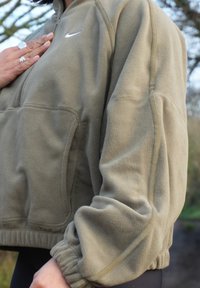 Personne portant une polaire Nike vert olive, la main posée sur la poitrine en plein air, avec des arbres et le ciel visibles en arrière-plan.