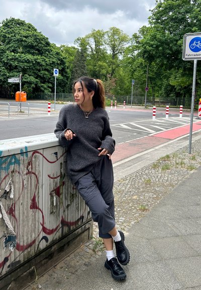 Suéter gris de gran tamaño, pantalones cortos grises y zapatos de plataforma negros. Calcetines blancos, collares en capas y peinado oscuro. Graffiti en la superficie cercana.