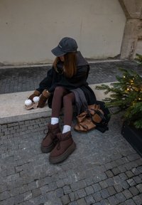 Femme en vêtements d'hiver assise sur un rebord en pierre, tenant deux tasses de café, avec un sac marron et un petit sapin décoré à proximité.