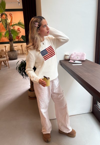 Joven con un clip de mariposa en el cabello, vistiendo un suéter blanco con la bandera americana, sosteniendo una taza amarilla, de pie junto al alféizar de una ventana en un café.