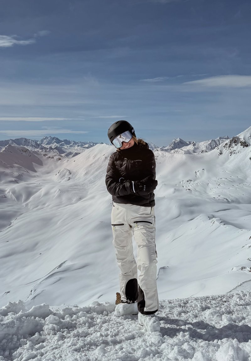 Persona con chaqueta negra, pantalones de esquí blancos, casco y gafas de esquí, de pie con los brazos cruzados en la cima de una montaña nevada bajo un cielo despejado.