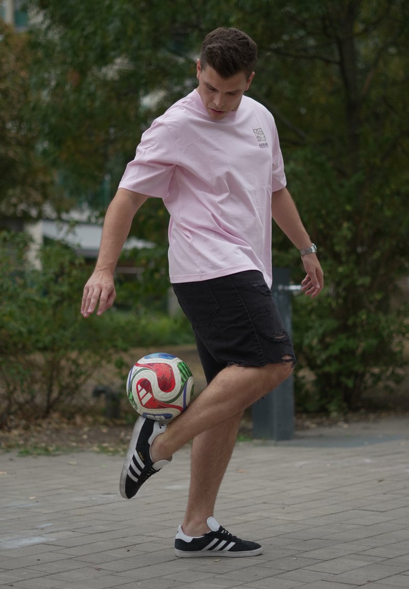 Un homme en chemise rose et short noir équilibre un ballon de football coloré sur son pied levé sur une surface pavée avec des arbres en arrière-plan.