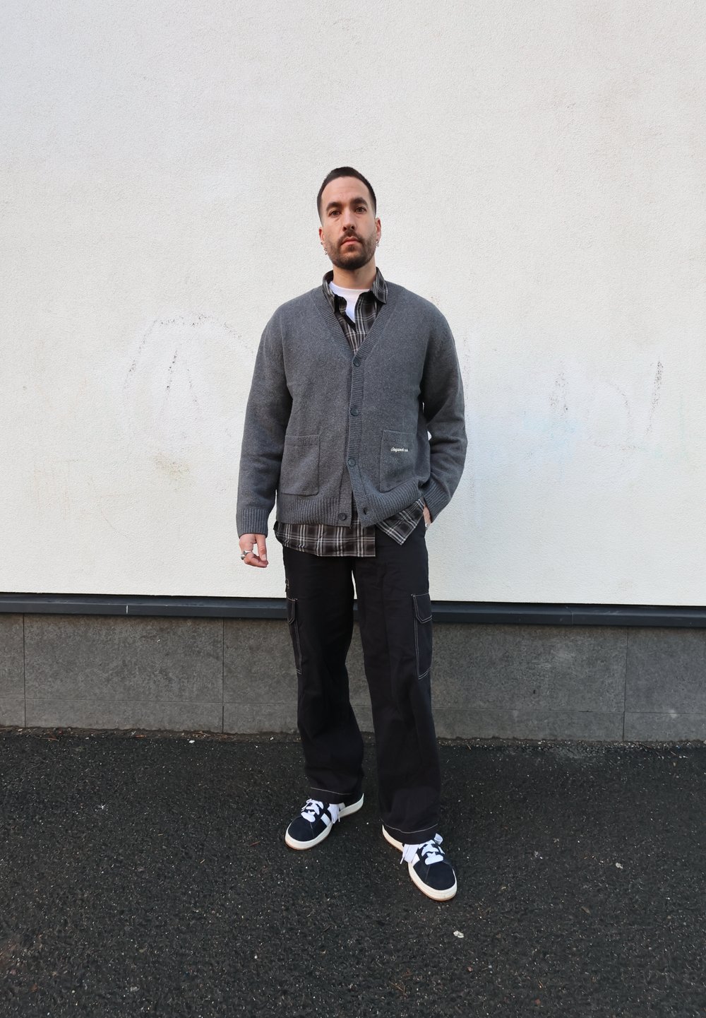 Cardigan en maille grise avec deux poches avant, porté sur une chemise à carreaux noire et blanche. Pantalon cargo noir et baskets complètent la tenue.