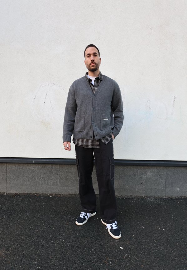 Grå strikket cardigan med to fremre lommer, brukt over en svart og hvit rutete skjorte. Sorte cargobukser og joggesko fullfører antrekket.