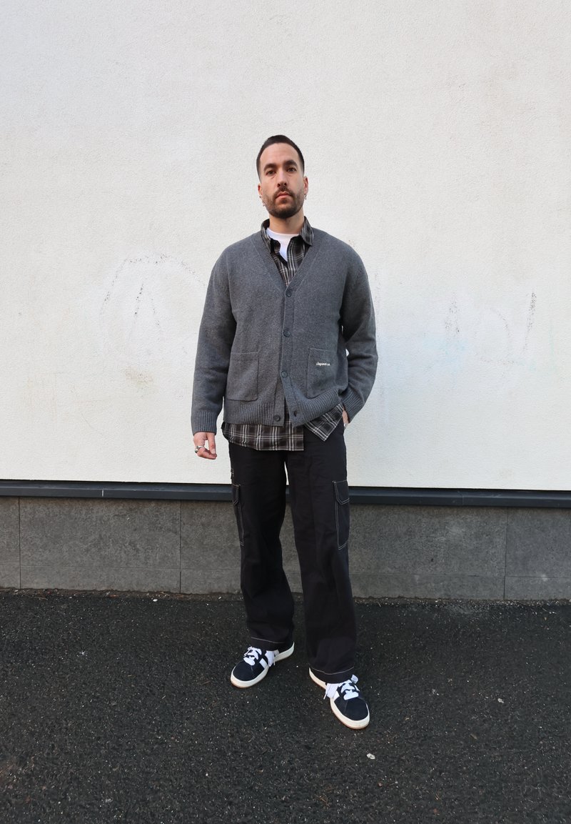Szürke kötött kardigán két első zsebbel, fekete-fehér kockás ing fölött viselve. A szettet fekete cargo nadrág és sportcipők egészítik ki.