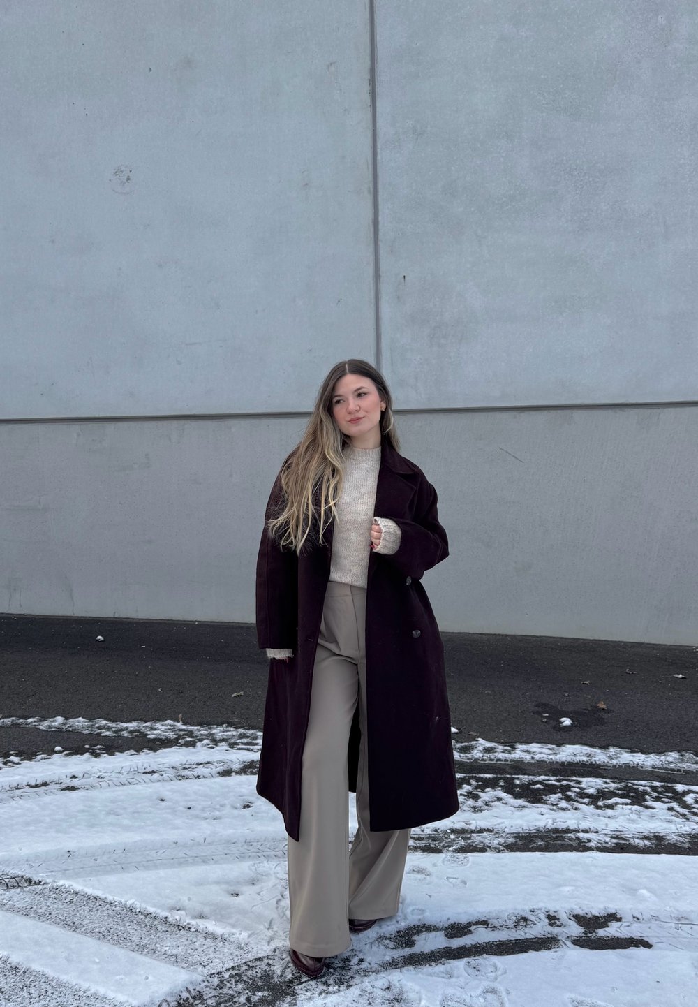 Νεαρή γυναίκα με μακριά μαλλιά φοράει μαύρο μακρύ παλτό, ανοιχτόχρωμο πουλόβερ και μπεζ παντελόνι φαρδύ στο κάτω μέρος, στέκεται σε χιονισμένο έδαφος δίπλα σε γκρίζο τοίχο.