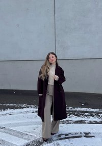 Νεαρή γυναίκα με μακριά μαλλιά φοράει μαύρο μακρύ παλτό, ανοιχτόχρωμο πουλόβερ και μπεζ παντελόνι φαρδύ στο κάτω μέρος, στέκεται σε χιονισμένο έδαφος δίπλα σε γκρίζο τοίχο.