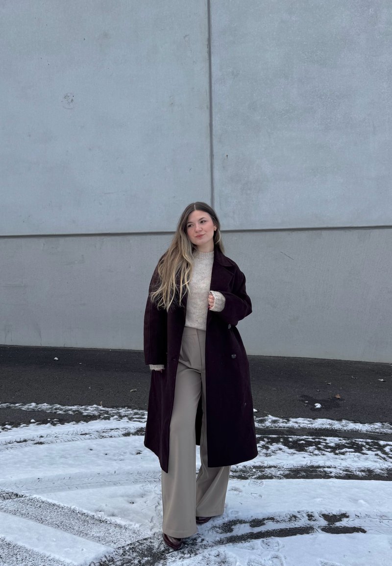 Tânără cu păr lung, purtând o haină lungă de culoare închisă, un pulover deschis la culoare și pantaloni largi bej, stând pe teren acoperit cu zăpadă lângă un perete gri.