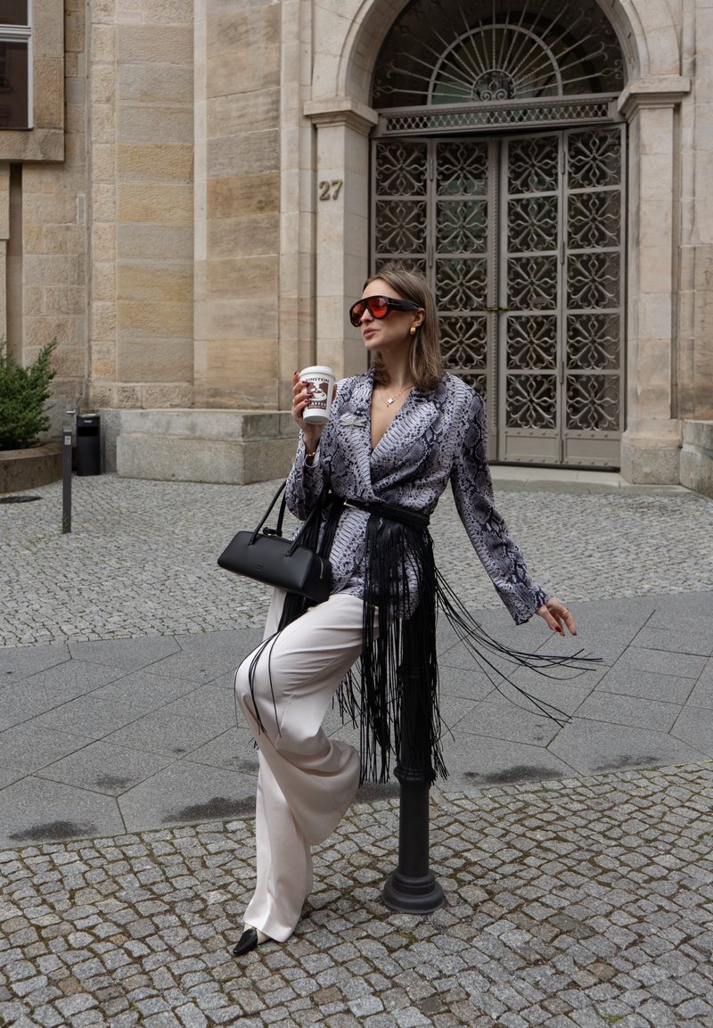 Femme vêtue d'une veste à motif serpent et d'un pantalon blanc, tenant une tasse de café, appuyée contre un poteau devant un bâtiment en pierre orné avec une grande grille métallique.