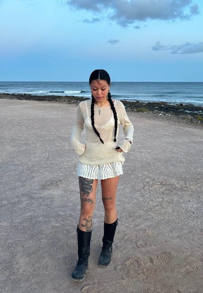 Top de manga larga transparente en crema con un diseño ajustado, combinado con una falda blanca de ganchillo. Botas de caña alta negras, tatuajes visibles en las piernas.