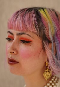 Profil af en person med pink ombre hår, lys orange øjenskygge, flere ansigtspiercinger og store guld hjerteformede øreringe.
