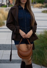 Casaco trench castanho sobre um macacão de denim, com uma mala de mão em pele castanha com um design curvo e alças finas. Em fundo paisagístico.