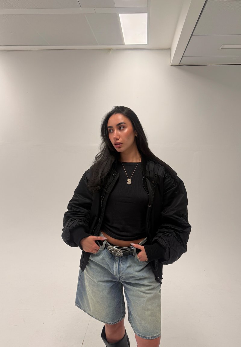 Frau mit langen dunklen Haaren, trägt eine schwarze Jacke, ein schwarzes Oberteil, Jeansshorts und einen Gürtel mit großer Schnalle, steht an einer weißen Wand.