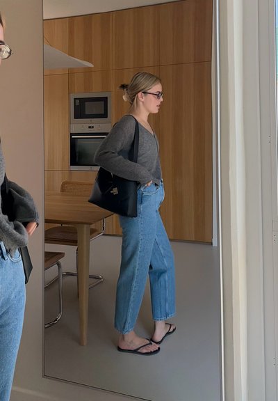 Mujer con gafas, suéter gris, vaqueros azules y sandalias negras, de pie en una cocina moderna con armarios de madera y una mesa de comedor.