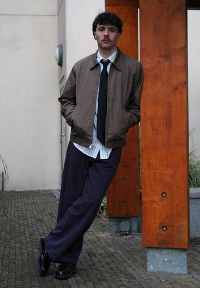 Chaqueta marrón con cremallera sobre una camisa de rayas blanca y azul, pantalones oscuros azules y zapatos negros brillantes, apoyado en un pilar de madera.