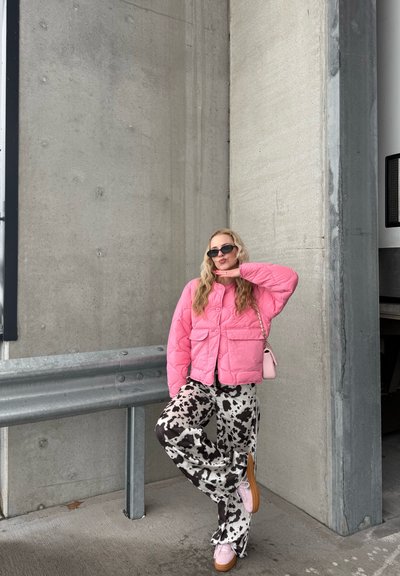 Mujer con chaqueta acolchada rosa, pantalones con estampado de vaca, zapatos rosas y gafas de sol, posando con un pie levantado contra una pared de concreto y una barandilla de metal.