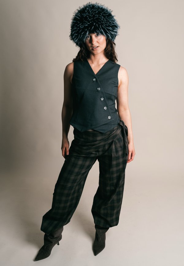 Femme portant un gilet sans manches sombre, un pantalon large à carreaux, des bottes à bout pointu et un grand chapeau noir duveteux, debout devant un fond uni.