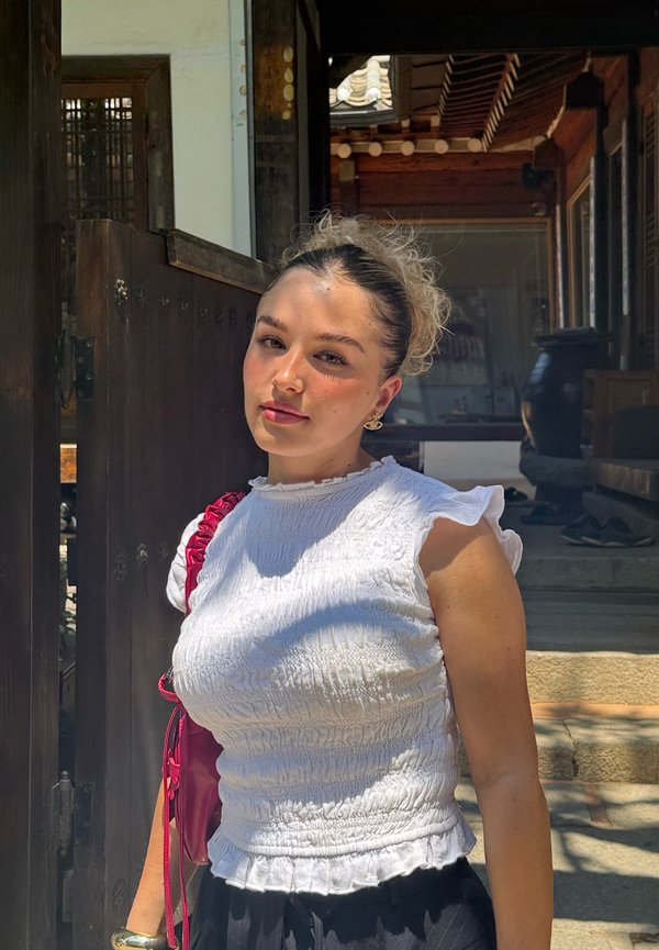 Giovane donna con capelli ricci che indossa una maglietta bianca con volant e una borsa rossa, in piedi nell'anticamera soleggiata di un edificio tradizionale in legno.