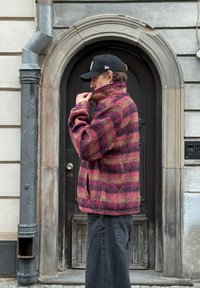 Veste en pullover duveteuse à rayures bordeaux et vertes, avec un col montant et des poches zippées, associée à un jean noir et une casquette.