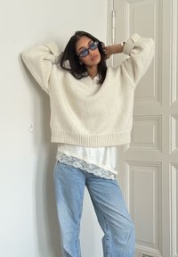 Pull crème oversize avec ourlet côtelé, associé à un haut blanc orné de dentelle et un jean bleu clair ample. Lunettes de soleil rondes portées.