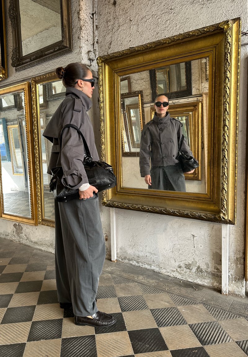 Mujer con chaqueta gris, pantalones anchos y gafas de sol, sosteniendo una bolsa negra, mirando su reflejo en un gran espejo adornado con un marco dorado ornamentado.