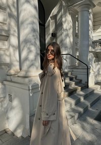 Μπεζ αδιάβροχο με ζώνη στη μέση, διπλές κουμπότρυπες και άνετη εφαρμογή. Στρογγυλά γυαλιά ηλίου συμπληρώνουν την κομψή εμφάνιση.