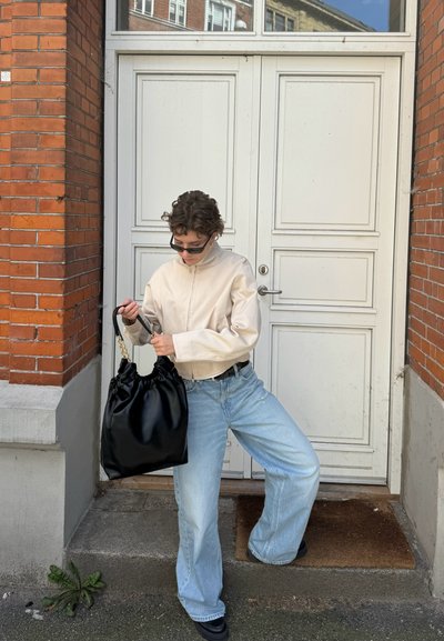 Chaqueta beige claro con cremallera, vaqueros de pierna ancha azules y un bolso de piel de charol negro con parte superior fruncida. Los accesorios incluyen gafas de sol oscuras.