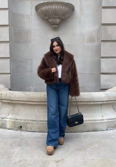 Chaqueta corta de piel sintética marrón, blusa blanca, jeans de talle alto, zapatos de plataforma en color tostado y un bolso negro acolchado. Materiales texturizados y corte de pierna ancha.