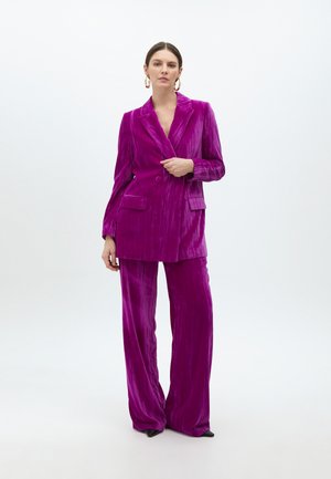 Rožu barvni žametni kostim, ki vključuje dvoredni blazer z reverji, stranskimi žepi in hlačami širokih nogavic. Tekstura je mehka z rahlim sijajem.