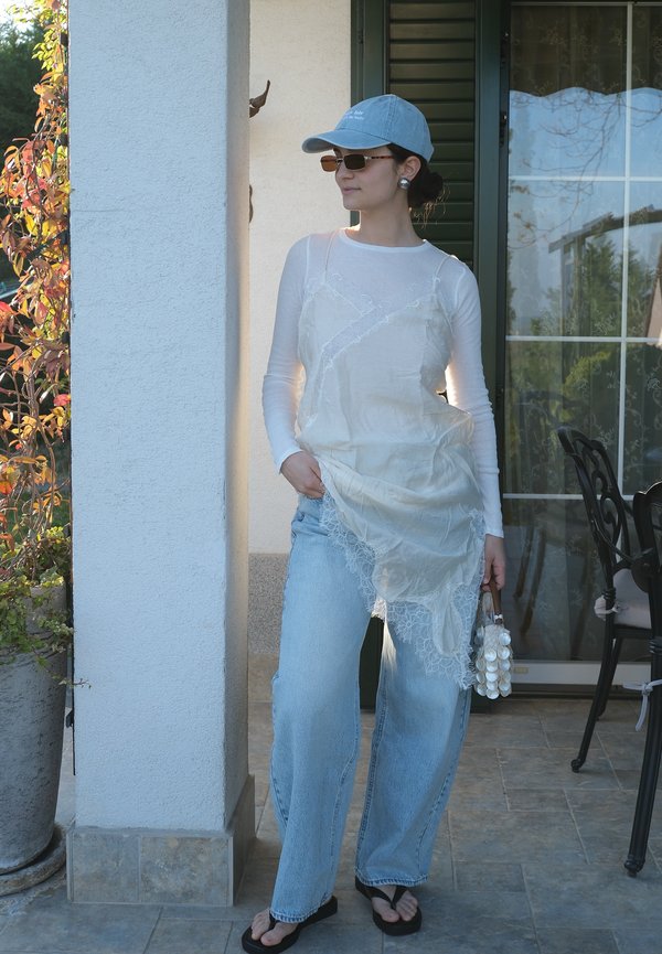 Žena nosiaca modrú čiapku, slnečné okuliare, priehľadné biele tričko s dlhým rukávom s čipkovanou spodničkou, svetlomodré džínsy, čierne sandále, drží bielu kabelku v tvare mušle.