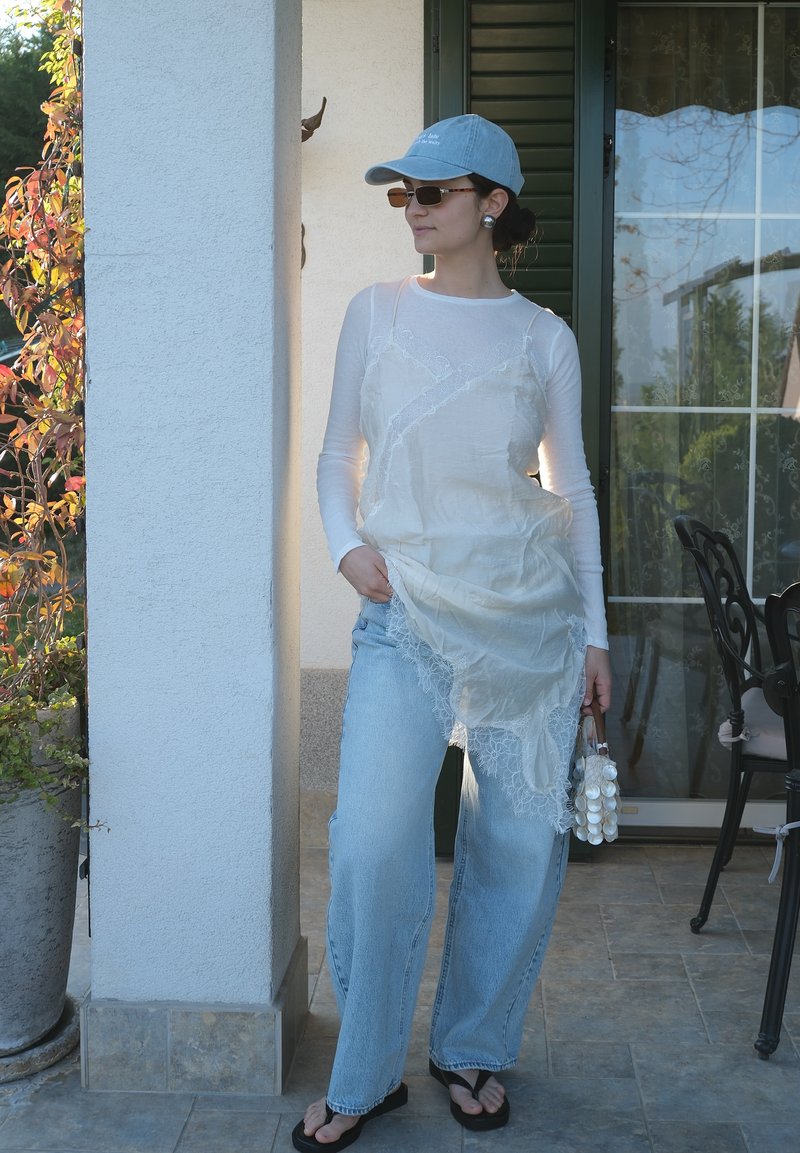 Γυναίκα με μπλε καπέλο, γυαλιά ηλίου, διάφανο λευκό μακρυμάνικο τοπ με δαντελένιο φόρεμα, ανοιχτό μπλε τζιν, μαύρα σανδάλια, κρατώντας μια λευκή τσάντα με σχέδιο κοχυλιού.