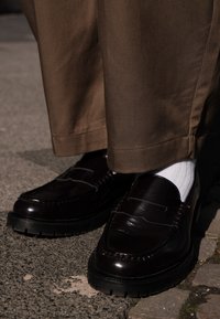 Mocassins de pele castanha com um acabamento brilhante, costura detalhada e uma sola de borracha robusta, combinados com meias brancas caneladas e calças bege.