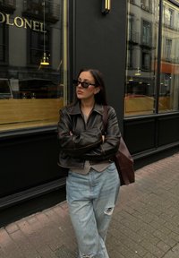 Must on must nahkajakk, millel on pruunid õmblused, hall särk all, sinised kulunud teksad ja pruun käekott, kandes tumedaid prille.