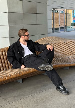 Hombre con chaqueta, pantalones y botas negras recostado en un banco de madera curvo frente a un edificio moderno con puertas de cristal.