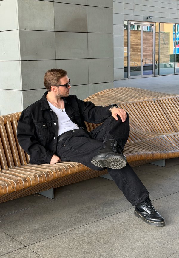 Mann mit schwarzer Jacke, Hose und Stiefeln lehnt sich auf einer gebogenen Holzbank vor einem modernen Gebäude mit Glastüren zurück.