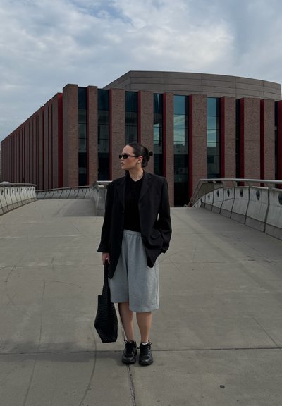 Mujer con blazer negro, pantalones cortos grises y zapatillas de deporte de pie en un puente de hormigón vacío con un edificio moderno de ladrillo y vidrio al fondo.