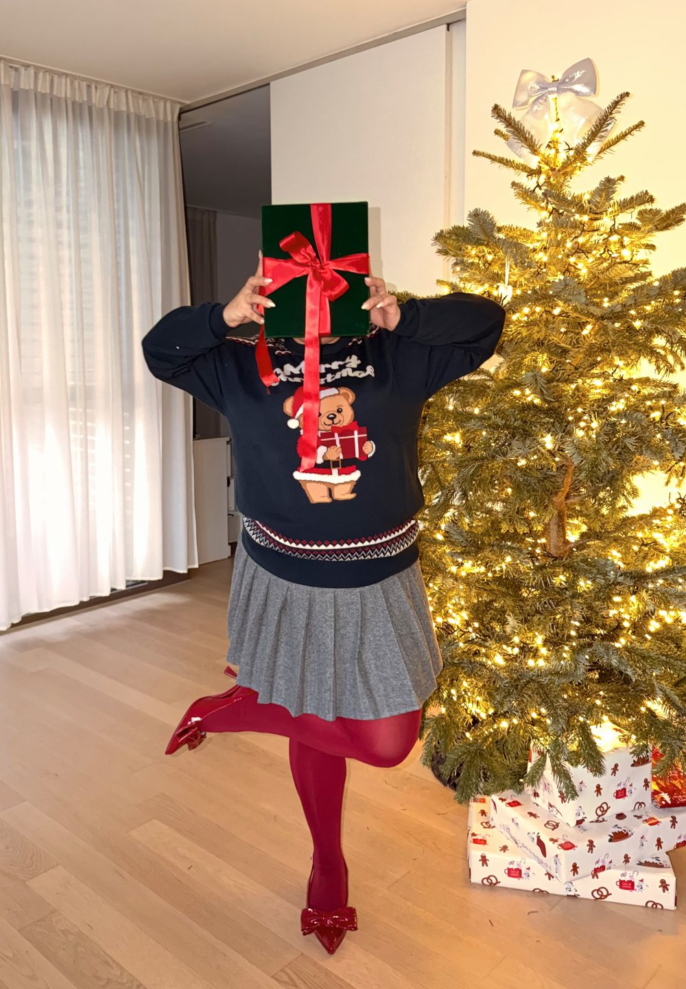 Scatola regalo verde con un nastro rosso tenuta davanti a una persona che indossa un maglione blu navy con un orso e una gonna grigia. Albero di Natale sullo sfondo.