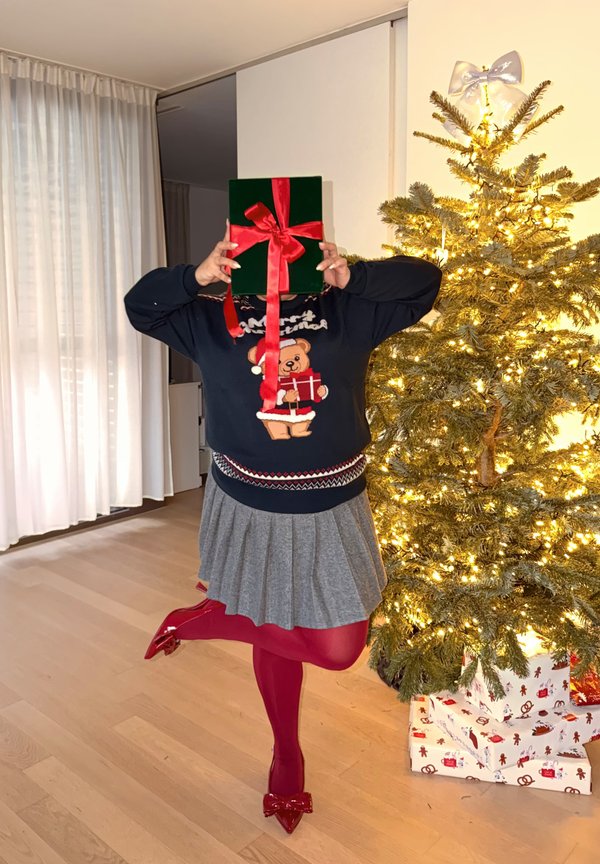 Groen geschenkdoosje met een rood lint, vastgehouden door een persoon die een marineblauwe trui met een beer draagt en een grijze rok. Kerstboom op de achtergrond.