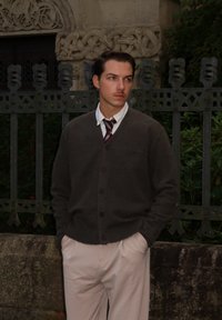Brauner gestrickter Cardigan über einem weißen Hemd mit einer Krawatte, hellbeige Hosen, stehend vor einem dekorativen schmiedeeisernen Zaun.