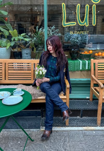 Mujer sentada en un banco de madera sosteniendo un ramo de tulipanes. Lleva un blazer oscuro y una blusa de cuadros, con jeans oscuros y tacones marrones. Decoración de café en tonos verdes.