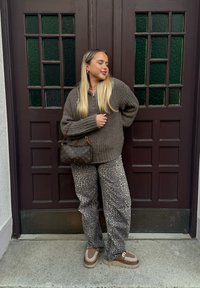 Γυναίκα με μακριά ξανθά μαλλιά, φορώντας oversized καφέ πουλόβερ, παντελόνι λεοπάρ, καφέ παπούτσια, κρατώντας μια καφέ designer τσάντα, στέκεται μπροστά από μια σκοτεινή πόρτα.